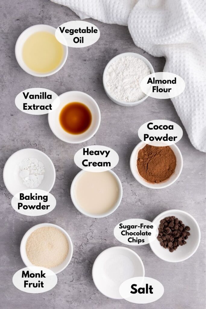 Nine labeled ingredients for a low carb microwave chocolate mug cake are arranged in small bowls on a gray surface: vegetable oil, almond flour, vanilla extract, cocoa powder, heavy cream, baking powder, sugar-free chocolate chips, monk fruit sweetener, and salt.