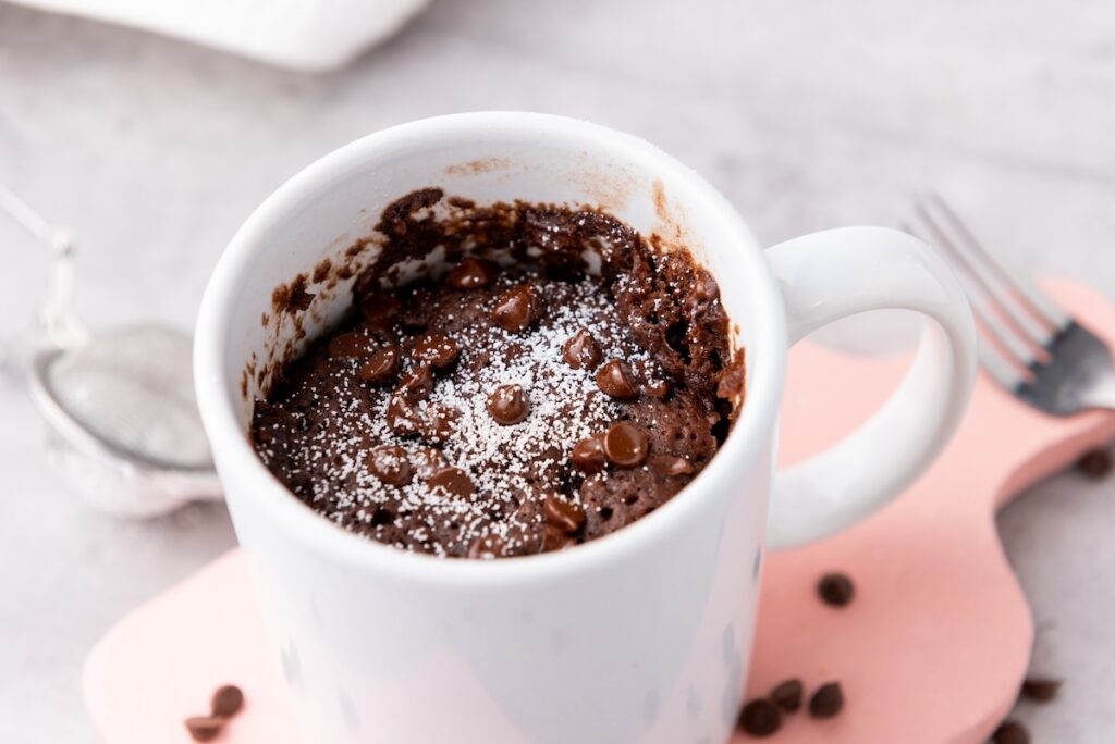 White mug with chocolate cake topped with chocolate chips and powdered sugar, on a pink surface. Fork and tea strainer are nearby.