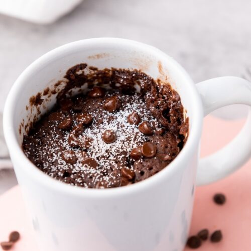 White mug with chocolate cake topped with chocolate chips and powdered sugar, on a pink surface. Fork and tea strainer are nearby.