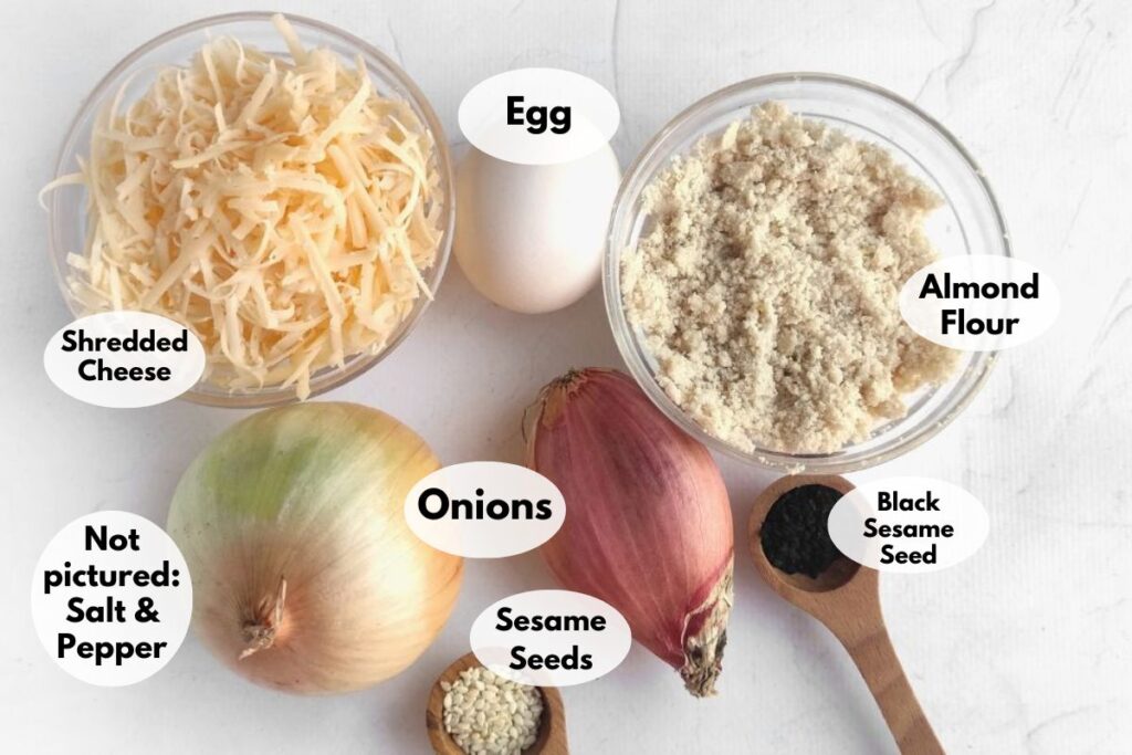 Ingredients on a white surface: egg, shredded cheese, almond flour, onion, shallot, black sesame seeds in a spoon. Not pictured: salt and pepper.