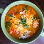 A green bowl filled with soup, topped with shredded cheese, sliced green onions, and pieces of red and green vegetables on a wooden surface.