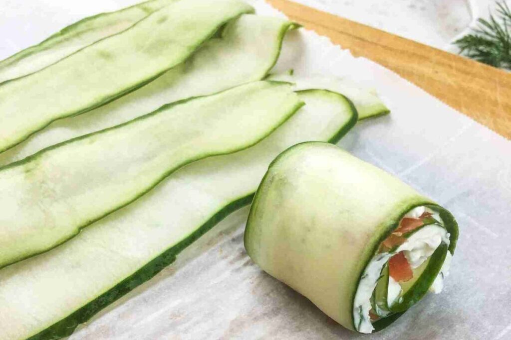 Thin slices of cucumber with one rolled into a spiral, filled with cream cheese and herbs, placed on a parchment paper.