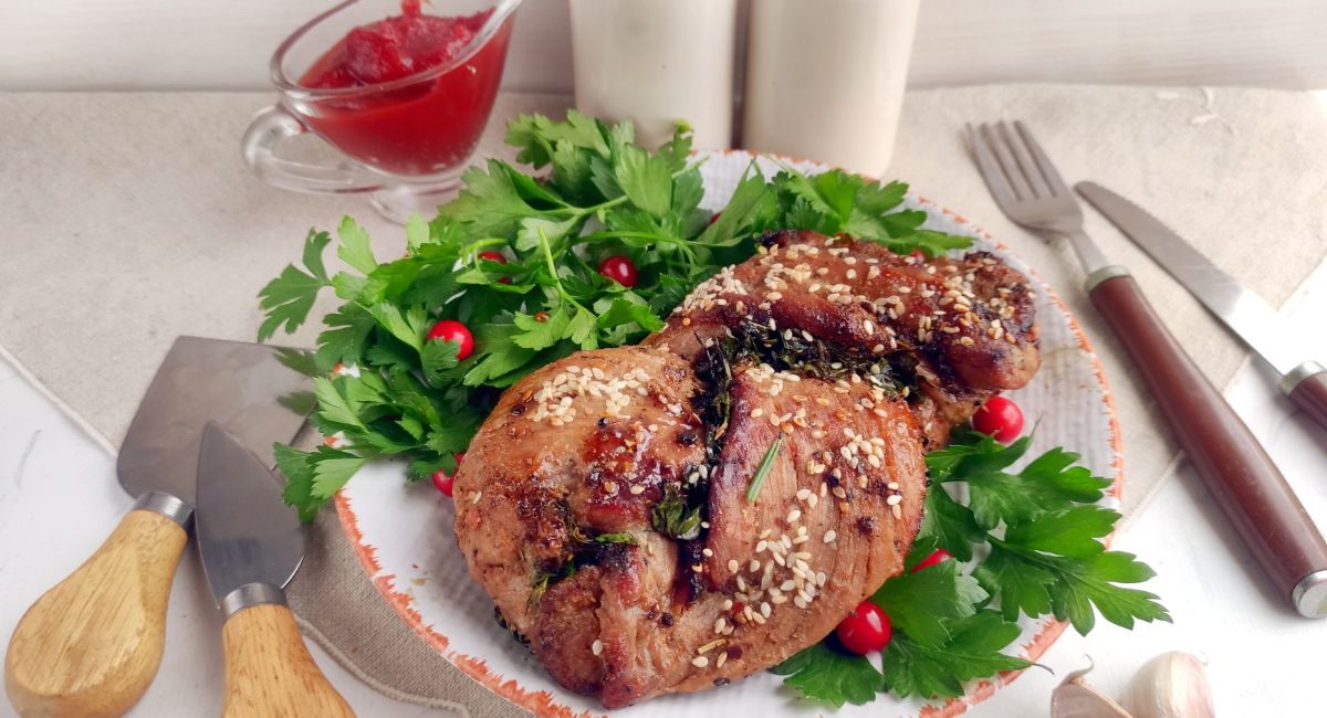 Cooked meat garnished with sesame seeds and herbs on a plate, surrounded by parsley and red berries, placed next to utensils and a small jug of sauce.