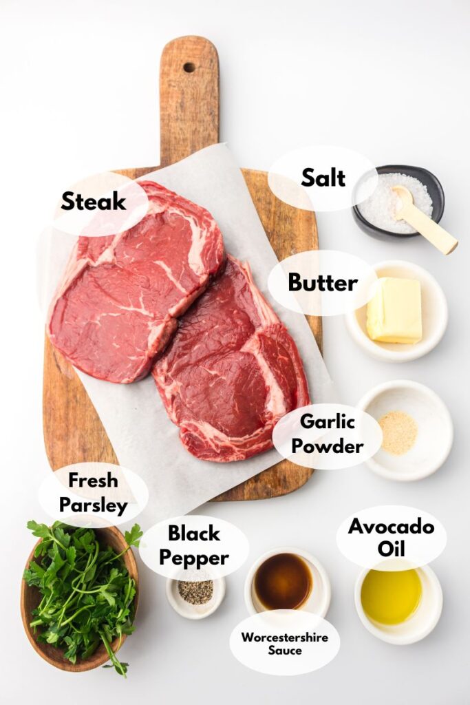 Raw steak on a cutting board, ready to transform into succulent air fryer steak bites, is surrounded by bowls of salt, butter, garlic powder, fresh parsley, black pepper, Worcestershire sauce, and avocado oil.