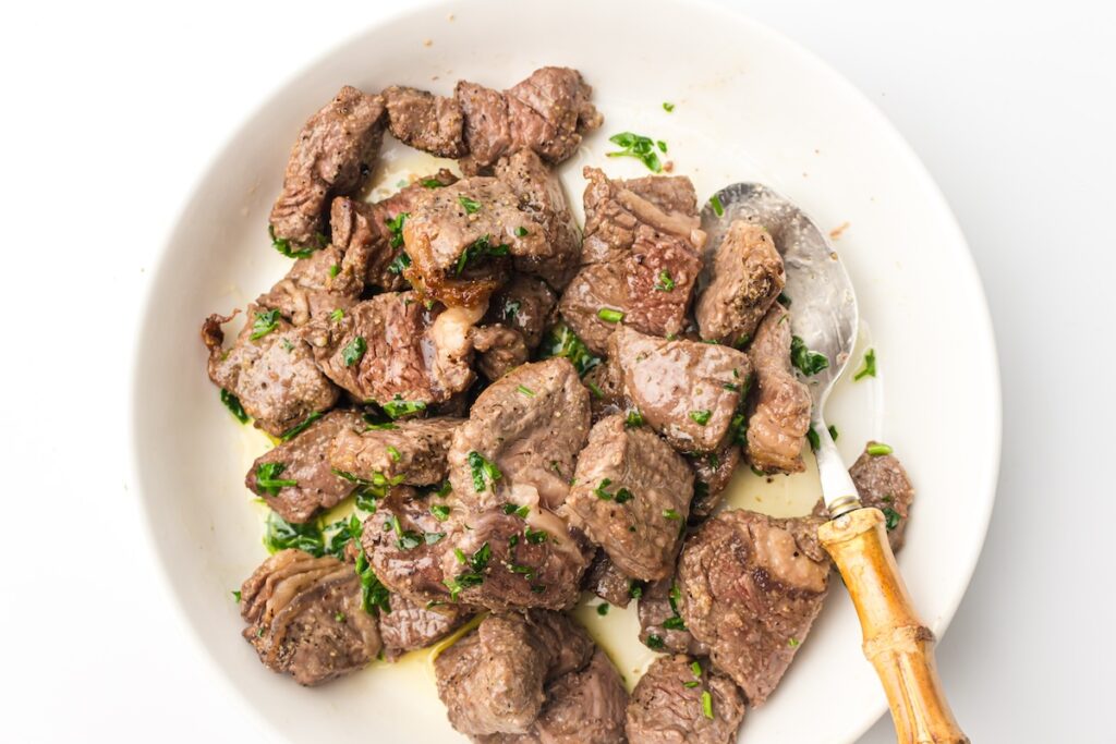 A white plate of air fryer steak bites, perfectly cooked in succulent chunks and garnished with parsley, served with a wooden-handled spoon.