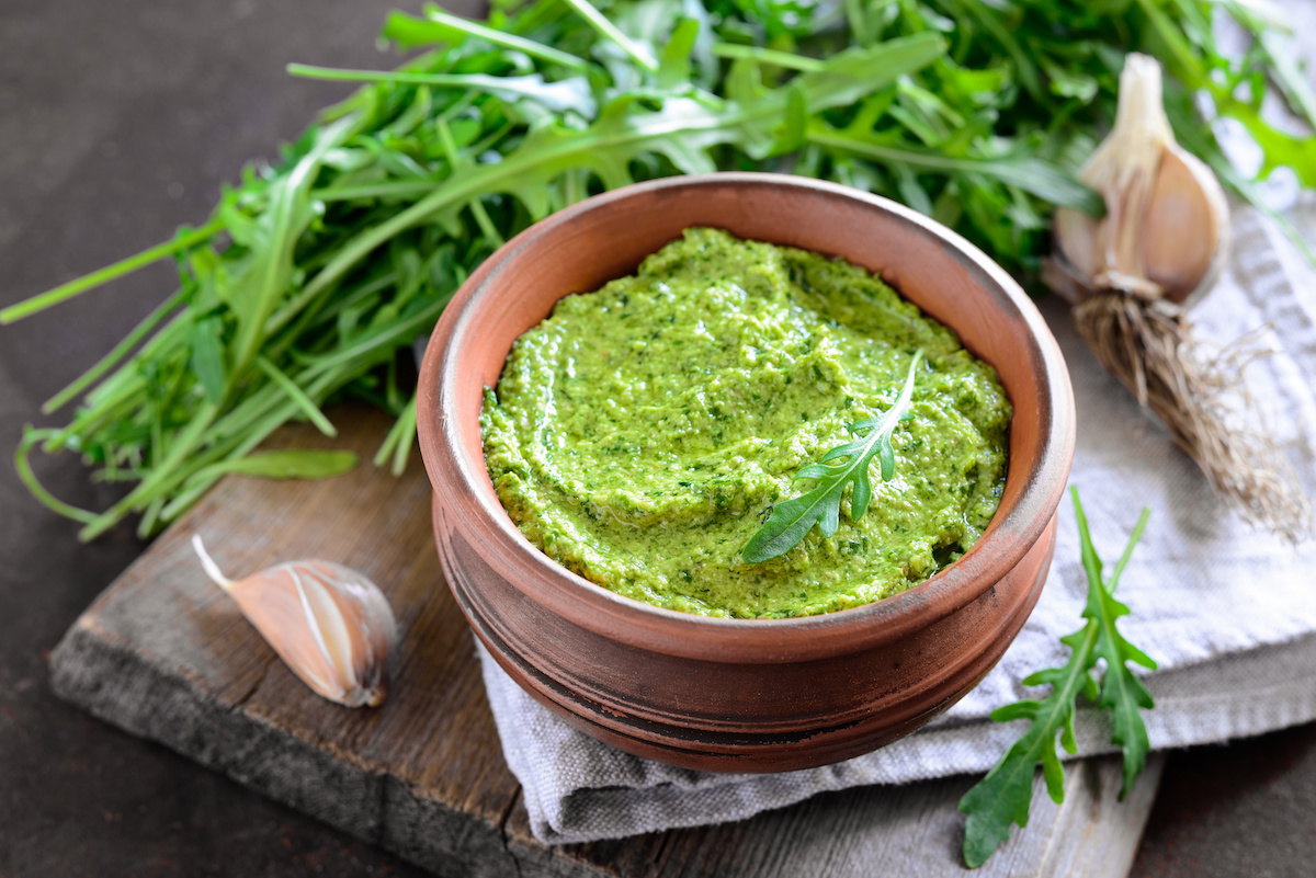 A wooden bowl of green pesto garnished with arugula, surrounded by fresh arugula leaves and garlic cloves on a rustic wooden surface.