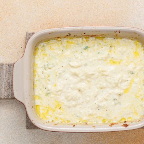 Baked cheesy dip in a rectangular dish placed on a stone board with a leather loop handle, showing a creamy texture with browned edges.