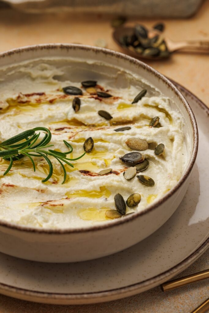 A bowl of creamy dip topped with olive oil, roasted seeds, and a sprig of rosemary on a plate with a fork in the background.