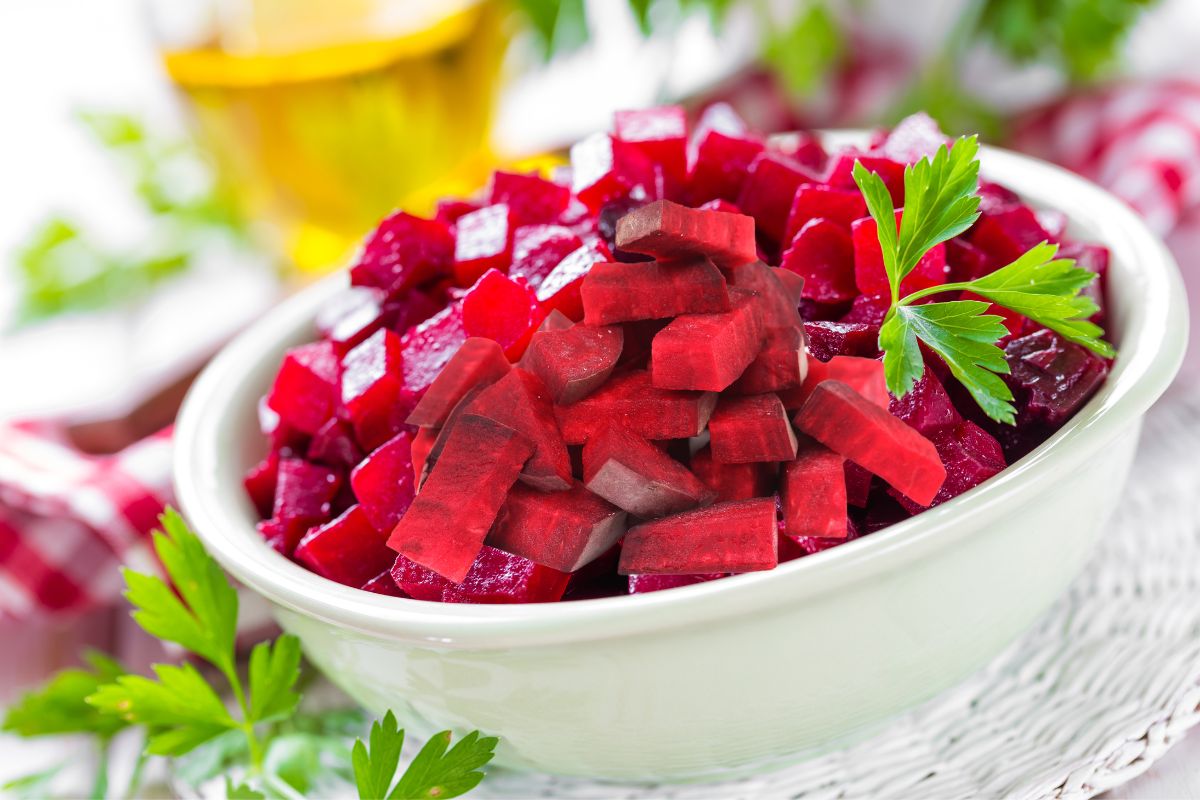 A bowl of diced beets garnished with parsley, set on a table with a blurred background of oil and checkered fabric.
