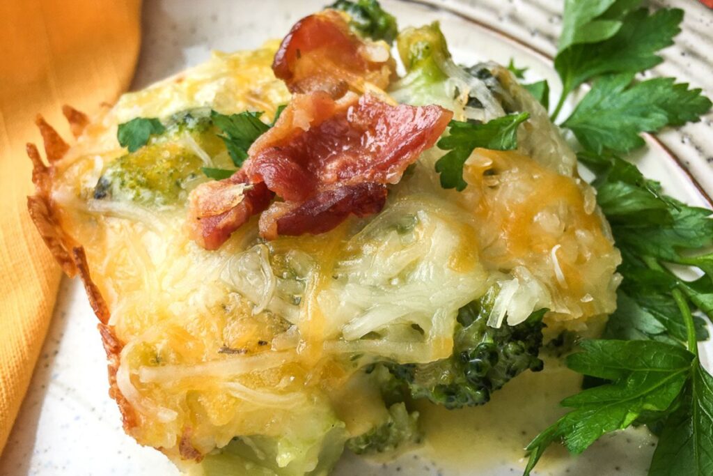 Broccoli casserole topped with melted cheese and crispy bacon, garnished with fresh parsley on a white plate.