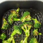 Cooked broccoli florets in an air fryer basket, lightly charred with visible seasoning specks.
