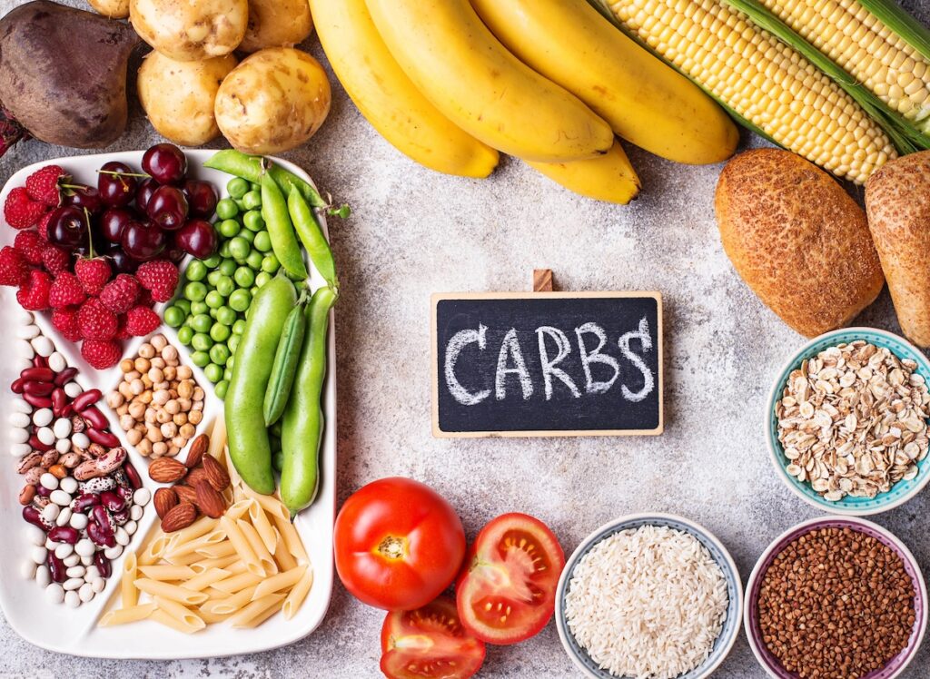 Assorted carbohydrate-rich foods, such as fruits, vegetables, grains, and pasta, are displayed alongside a small chalkboard labeled "CARBS" on a textured surface—perfect for exploring net carbs versus total carbs.
