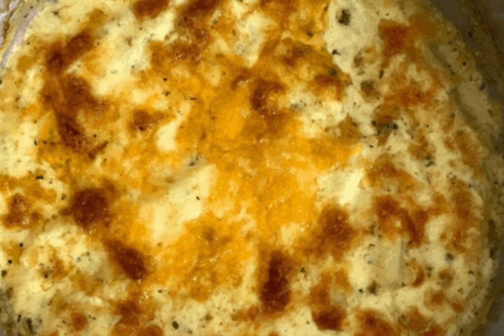 Close-up of a golden brown cheesy casserole, possibly containing potatoes and herbs, baked in a circular dish.