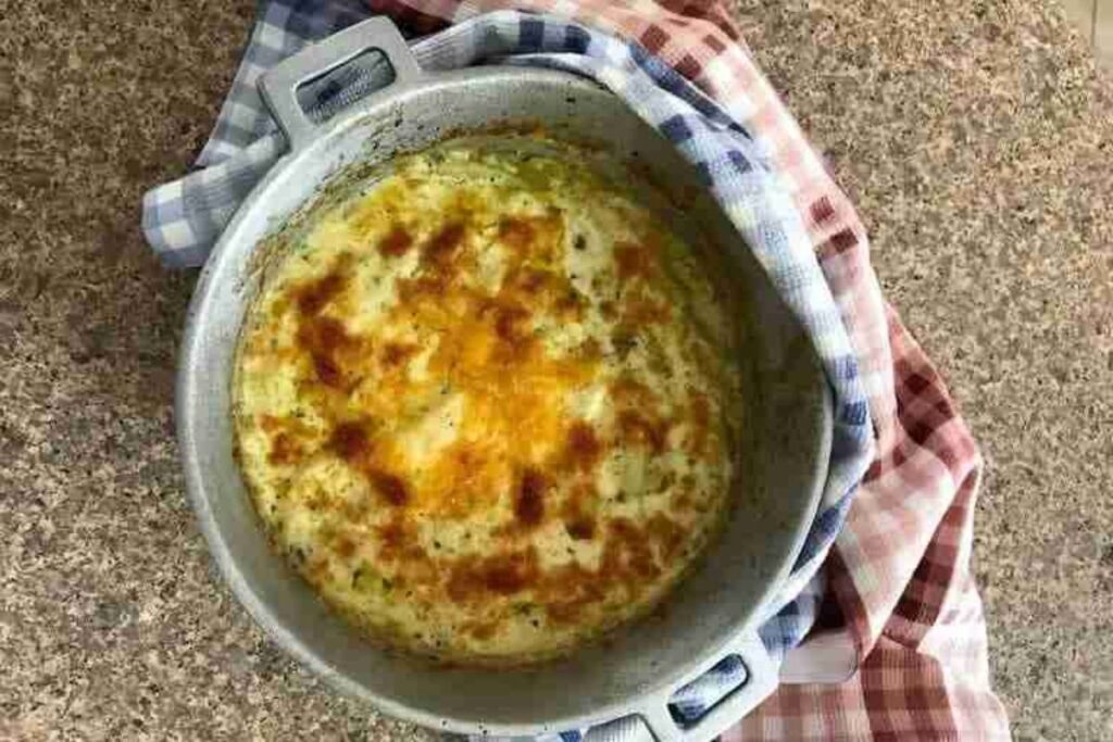 A baked gratin dish with a golden, bubbly top sits in a round pan. The pan is placed on a countertop, partially covered by a checkered cloth.