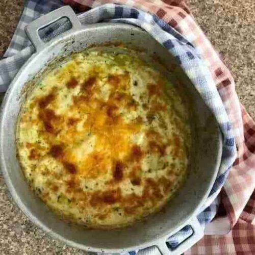 A baked gratin dish with a golden, bubbly top sits in a round pan. The pan is placed on a countertop, partially covered by a checkered cloth.