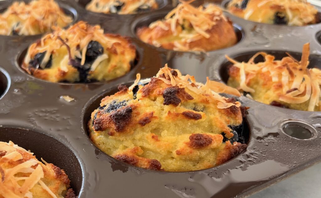 Close-up of freshly baked low carb blueberry muffins with a golden-brown top and toasted coconut flakes, nestled in a dark muffin tin.