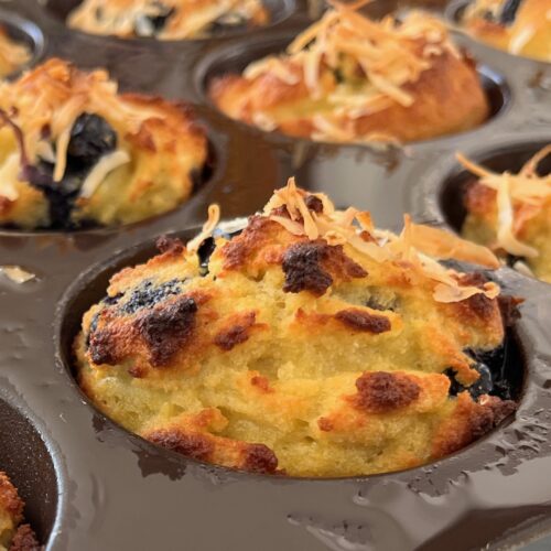 Close-up of freshly baked low carb blueberry muffins with a golden-brown top and toasted coconut flakes, nestled in a dark muffin tin.
