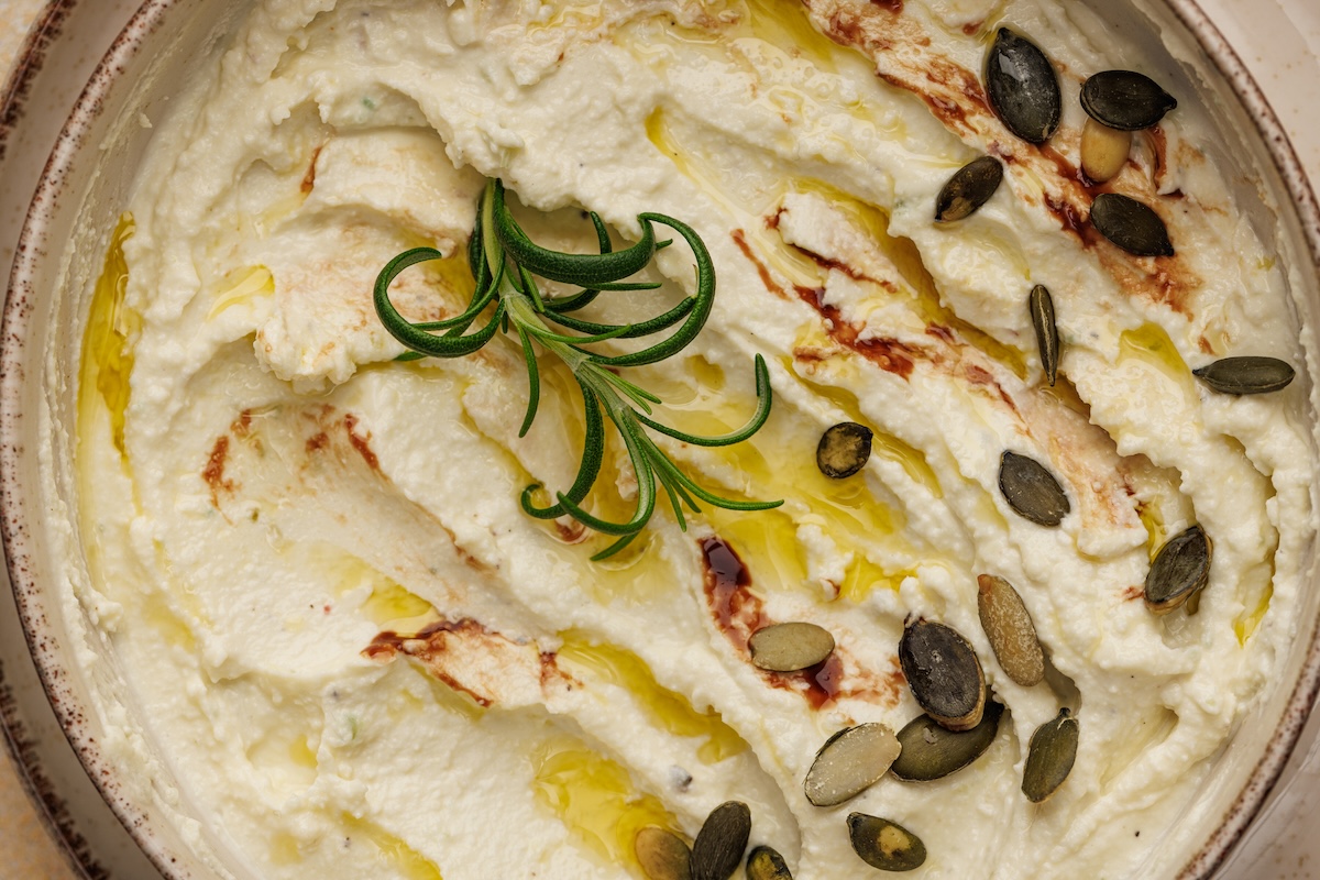A bowl of creamy dip garnished with a sprig of rosemary, a drizzle of oil, and pumpkin seeds.