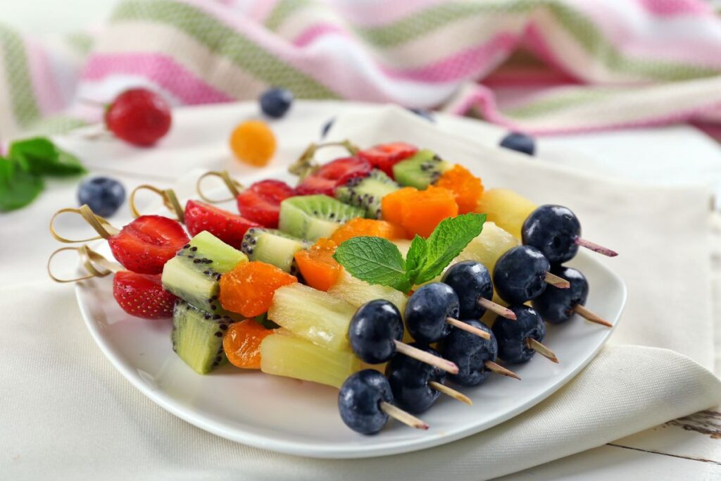 Fruit skewers with grapes, kiwi, pineapple, strawberries, and melon elegantly arranged on a white plate. These vibrant treats make for a delightful low-carb party food option, complemented by the charming striped cloth in the background.