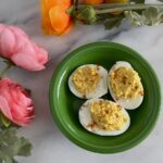 Three deviled eggs on a green plate with paprika seasoning. Surrounding are pink and orange artificial flowers on a marble surface.