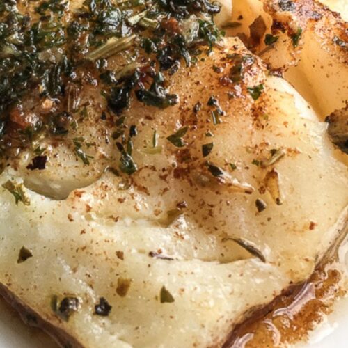 Close-up of a seasoned and cooked fish filet on a white plate, garnished with herbs and spices.