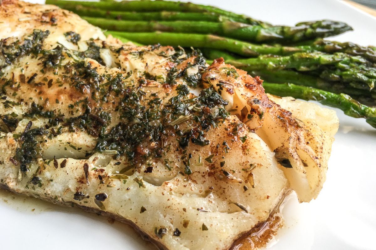 Grilled fish fillet with herbs served alongside cooked asparagus on a white plate.