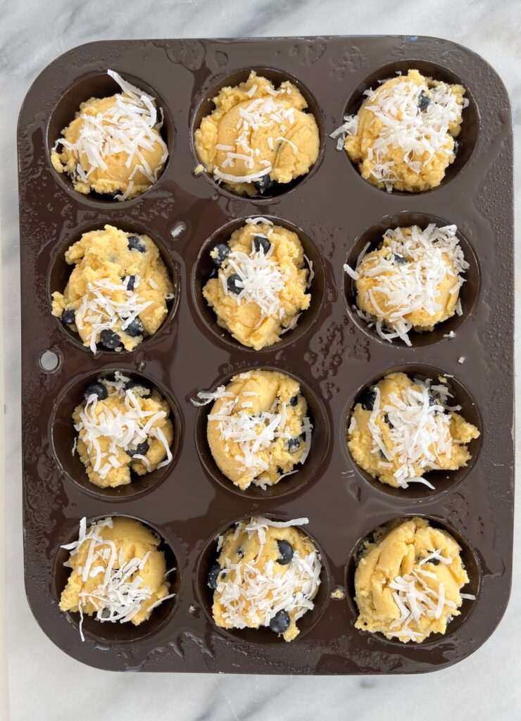 Low carb muffin tray filled with unbaked batter, rich in blueberries, crowned with a sprinkle of shredded coconut.