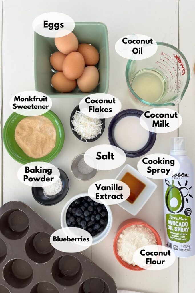 Ingredients for baking laid out: eggs, coconut oil, coconut milk, coconut flour, coconut flakes, monkfruit sweetener, baking powder, salt, vanilla extract, cooking spray, and blueberries—all set for delicious low carb blueberry muffins.
