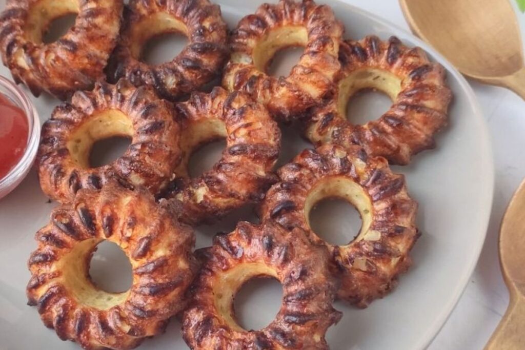 A plate of crispy, browned ring-shaped treats, resembling bundt cakes, next to wooden utensils and a small cup of red sauce.