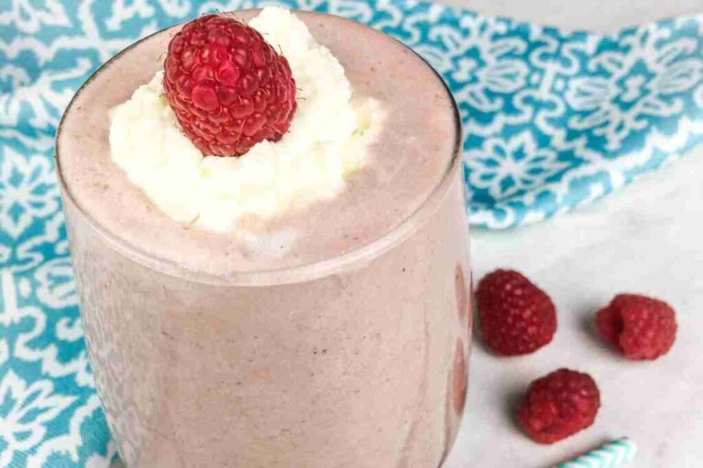 A low carb chocolate smoothie in a glass, topped with whipped cream and a raspberry, features three additional raspberries and a straw on the side, all set against a patterned blue cloth background.
