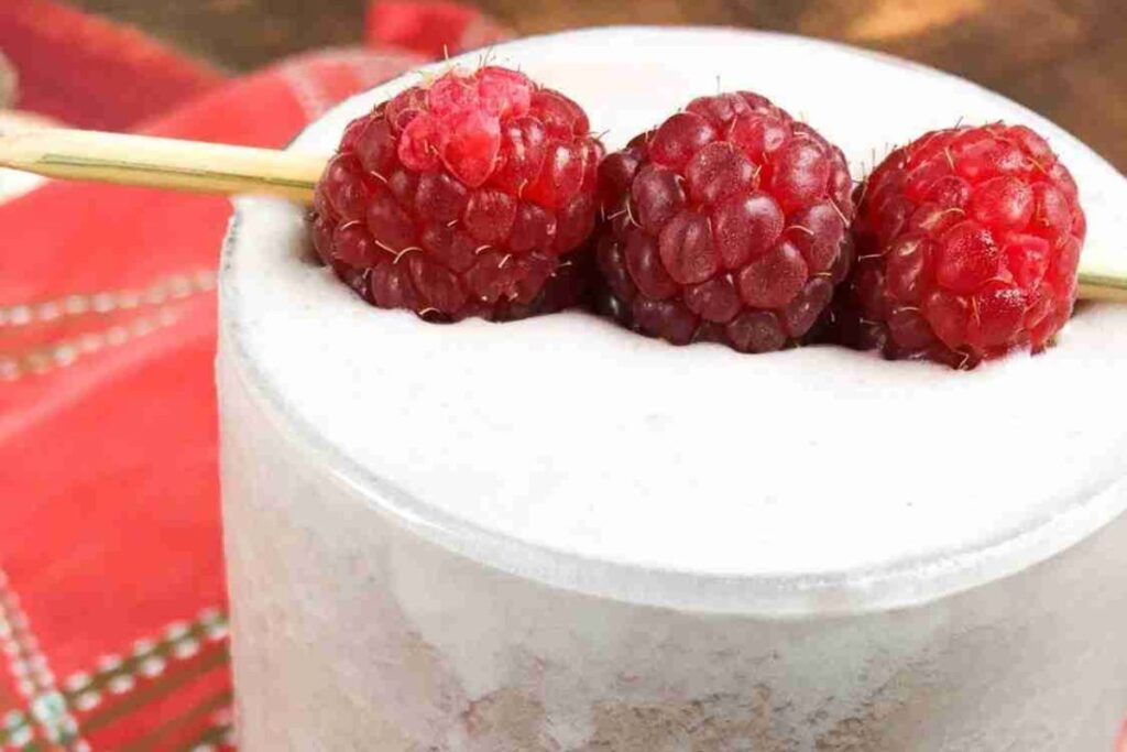 A low-carb raspberry smoothie, topped with frothy foam, features three raspberries on a skewer gracefully resting on the rim.