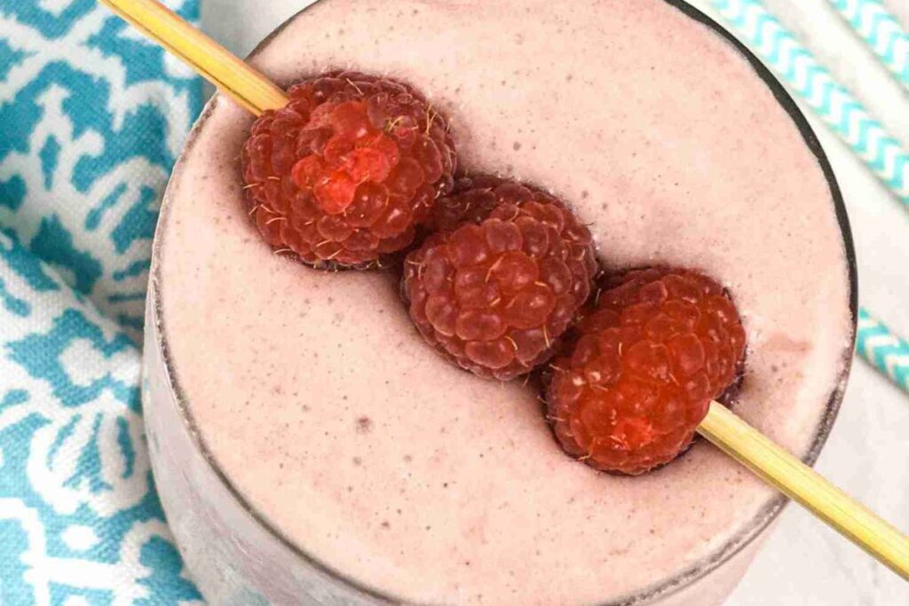 A low carb raspberry smoothie in a glass, crowned with three raspberries on a skewer. A blue patterned cloth peeks from the side.