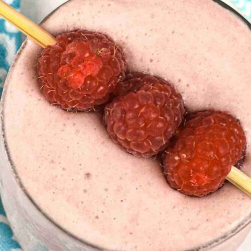 A low carb raspberry smoothie in a glass, crowned with three raspberries on a skewer. A blue patterned cloth peeks from the side.