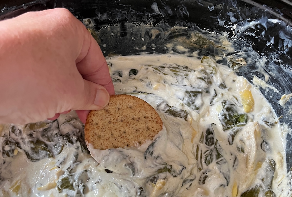 A hand holding a cracker dips it into a creamy spinach and artichoke mixture.