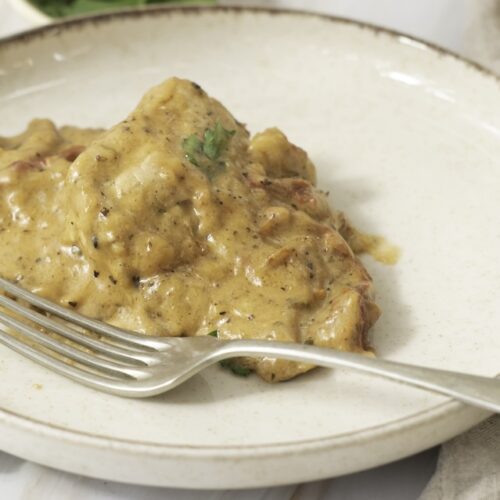 A plate with a serving of meat covered in creamy sauce, garnished with herbs. A fork rests on the plate.