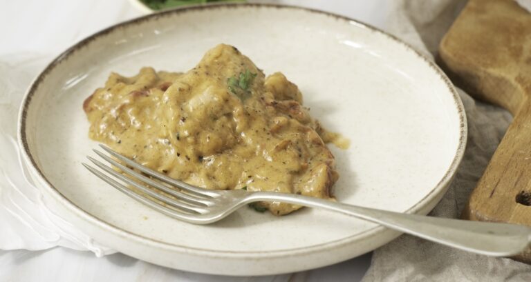 A plate with a serving of meat covered in creamy sauce, garnished with herbs. A fork rests on the plate.