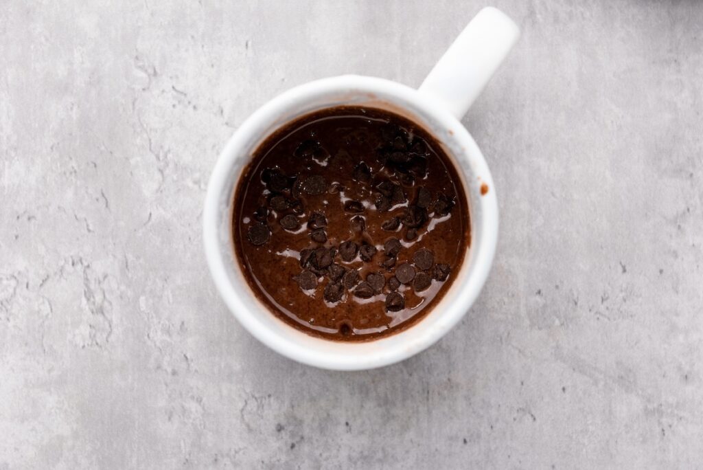 A mug filled with chocolate dessert topped with dark chocolate chips on a gray textured surface.