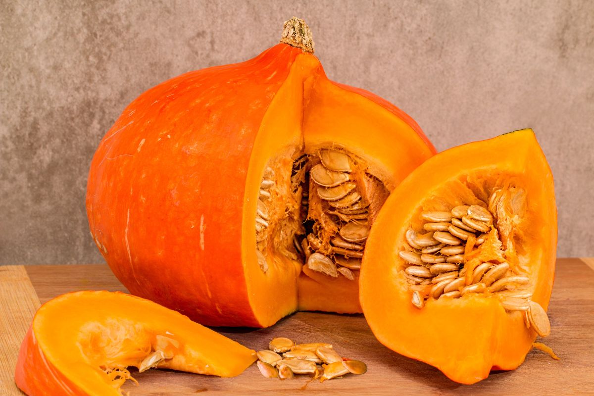 Orange pumpkin cut open, showing seeds and pulp inside, placed on a wooden surface with a gray textured background.