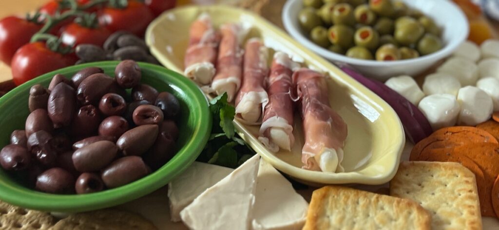 A platter brimming with low carb party food: olives, stuffed prosciutto, tomatoes, cheese, crackers, and mozzarella balls.