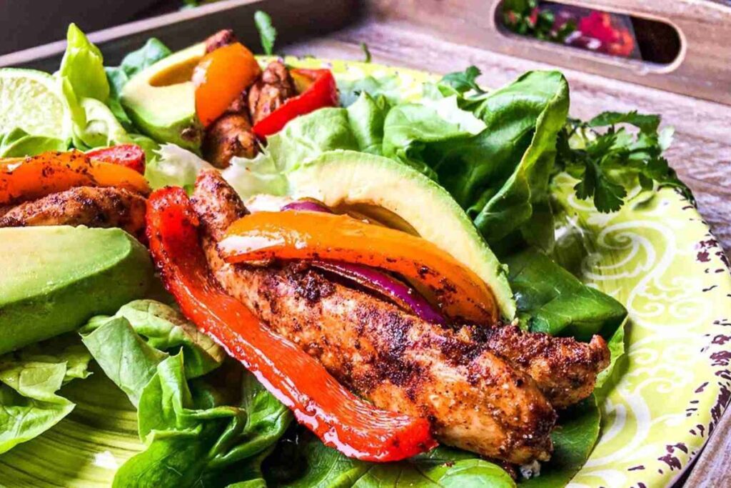 Grilled chicken strips mingle with red and yellow peppers, avocado, and lettuce on a green plate, reminiscent of vibrant sheet pan chicken fajitas.