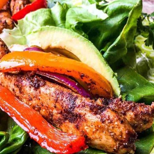 Grilled chicken strips with red and yellow peppers on lettuce, garnished with avocado slices, reminiscent of sheet pan chicken fajitas, artfully arranged on a decorative plate.