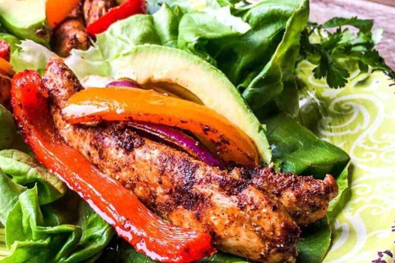 Grilled chicken strips with red and yellow peppers on lettuce, garnished with avocado slices, reminiscent of sheet pan chicken fajitas, artfully arranged on a decorative plate.