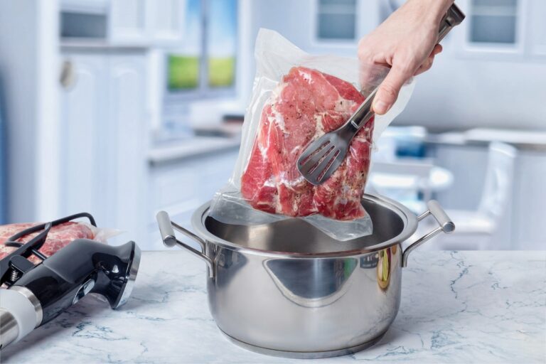 A hand lowers a vacuum-sealed bag of raw meat with herbs into a pot on a marble countertop, with a sous vide device nearby.