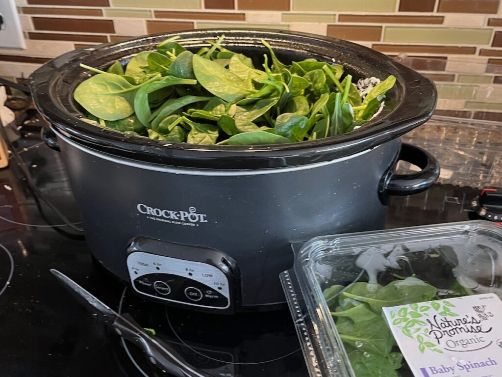 A slow cooker brims with fresh spinach on the kitchen counter, setting the stage for a delicious low carb spinach artichoke dip, while an open container of baby spinach sits invitingly nearby.