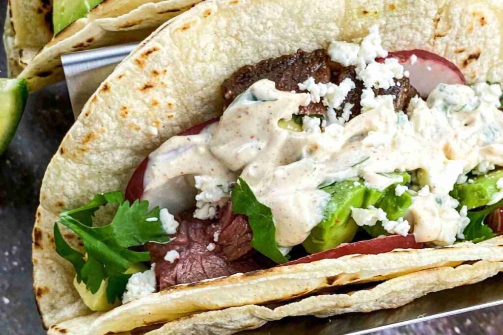 A taco filled with grilled steak, avocado, tomato slices, crumbled cheese, cilantro, and creamy sauce in a flour tortilla.