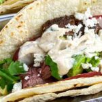 A taco filled with grilled steak, avocado, tomato slices, crumbled cheese, cilantro, and creamy sauce in a flour tortilla.