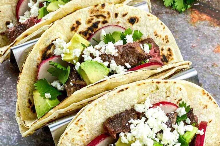 Three beef tacos with avocado, radish slices, cilantro, and crumbled cheese in taco holders on a textured surface.