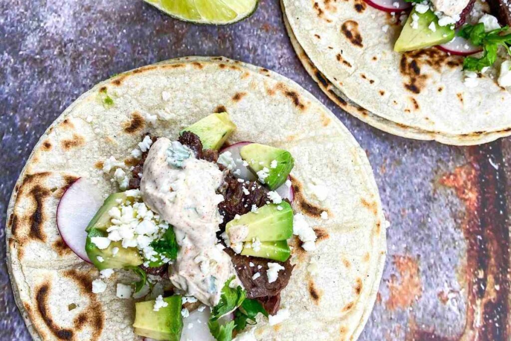 Two tortillas topped with beef, avocado, sliced radishes, crumbled cheese, and a creamy sauce. Lime wedge on the side.