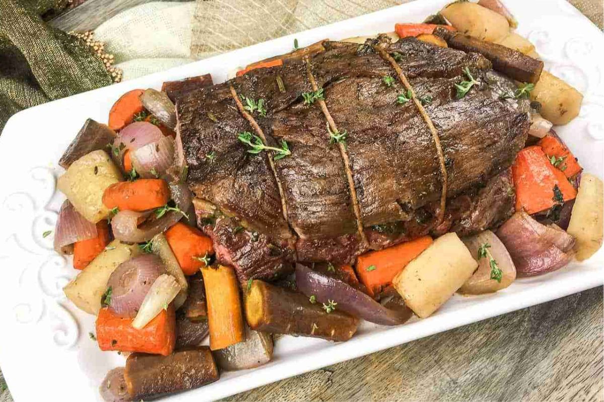 A roasted beef pot roast with twine, surrounded by cooked carrots, onions, and potatoes on a white platter.