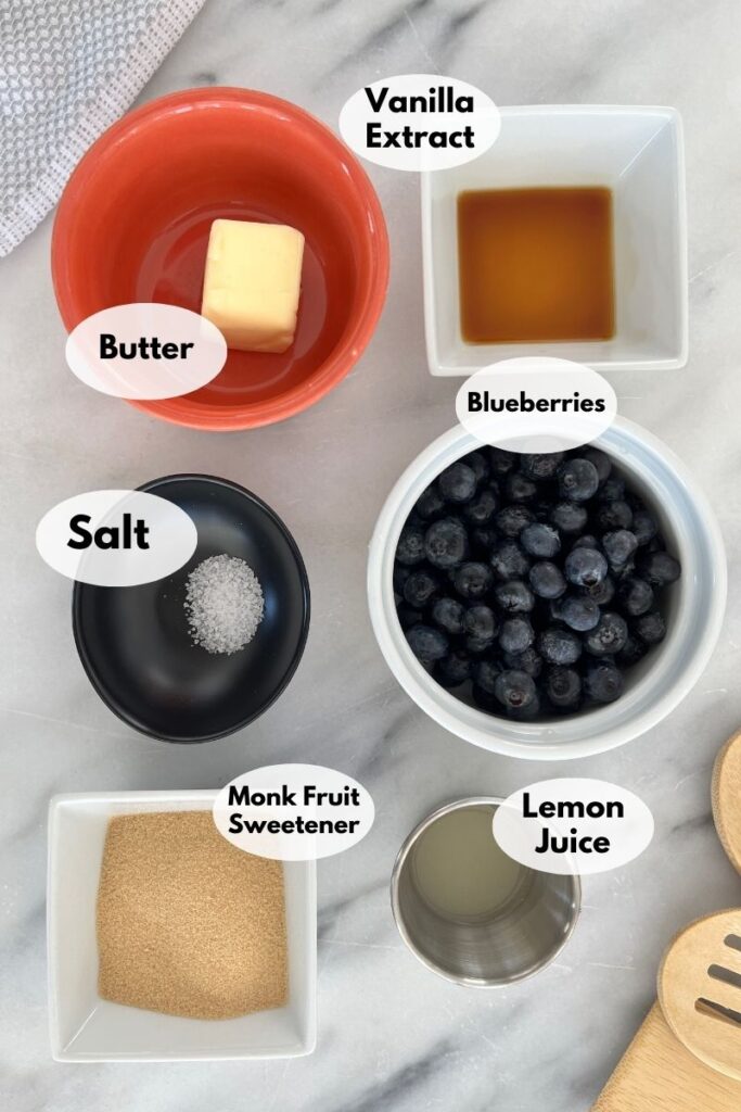 Ingredients for a sugar-free blueberry compote are displayed in separate bowls: butter, vanilla extract, salt, blueberries, monk fruit sweetener, and lemon juice.